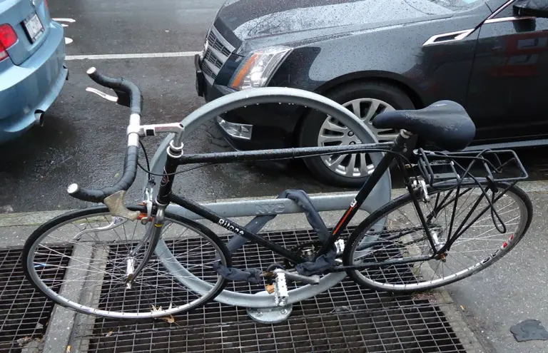 Bicicleta atada a un soporte fijo en la calle, con gente alrededor.