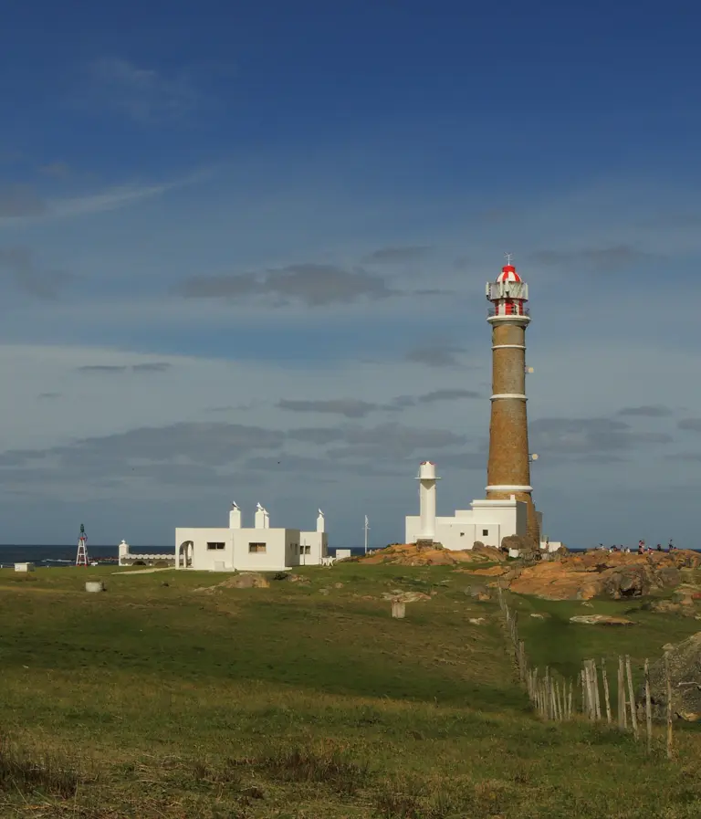 A stone lighthouse with white bands and a red-and-white lantern beside a smaller white tower and whitewashed houses on a grassy field.