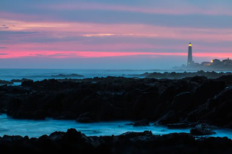 Dark rocks and long-exposure surf at dusk; far off, a lit lighthouse under a pink sky.