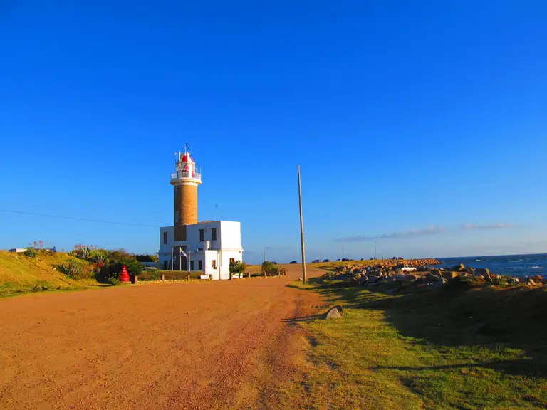 A brick lighthouse with a red lantern above a white building, beside a dirt track and a rocky shore.