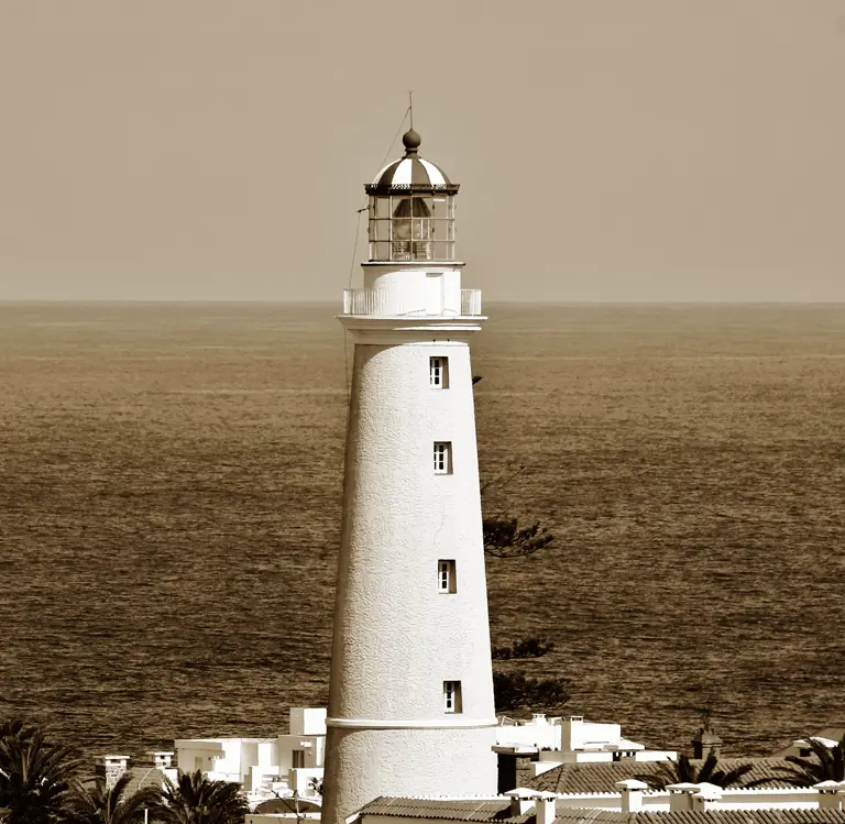 Sepia-toned photo of a white lighthouse with a striped lantern, above rooftops and palms, sea behind.