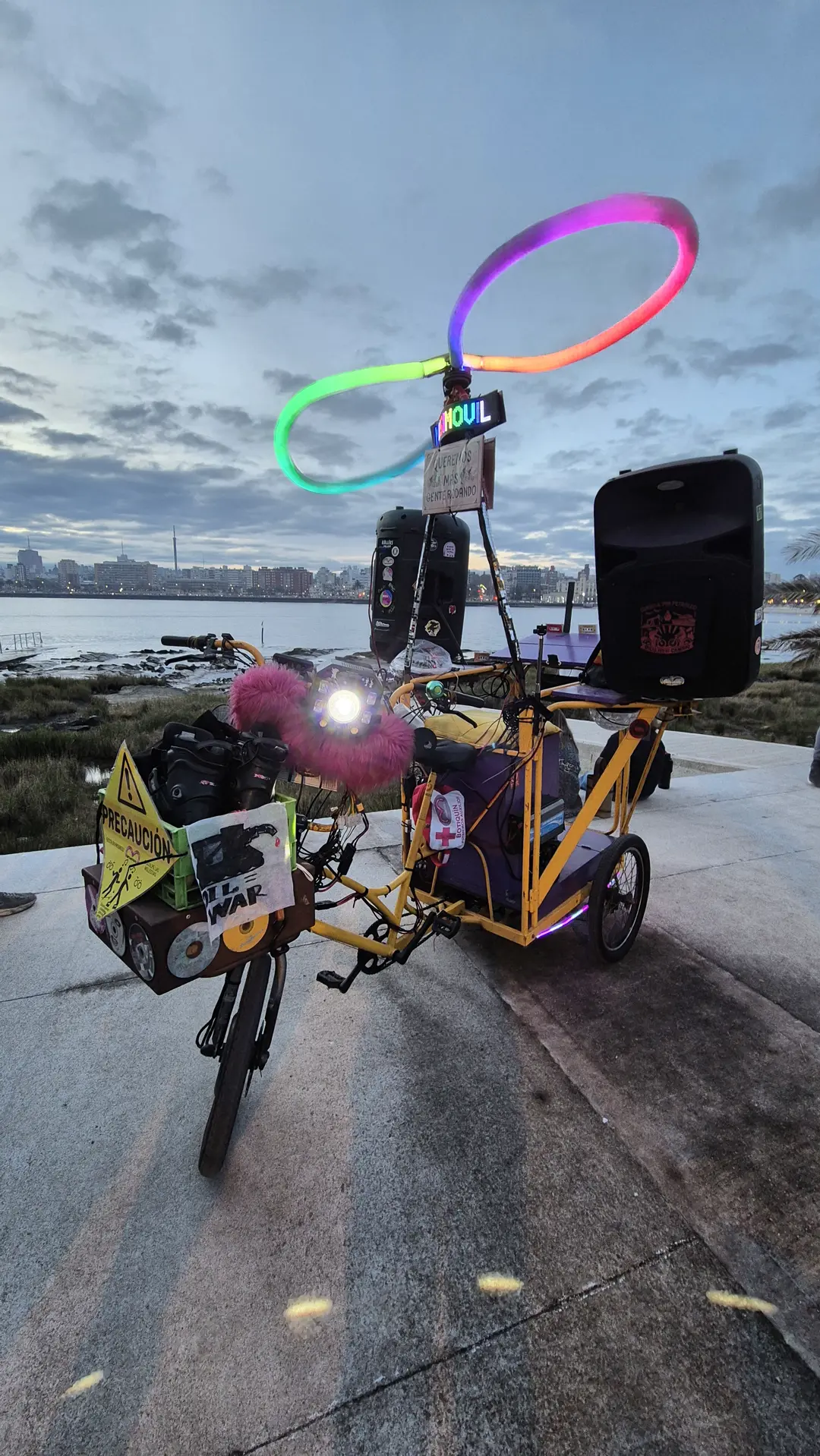 A pedal tricycle carrying a speaker stack, a curved neon hoop arcing above the load, alone on the paving beside the water.