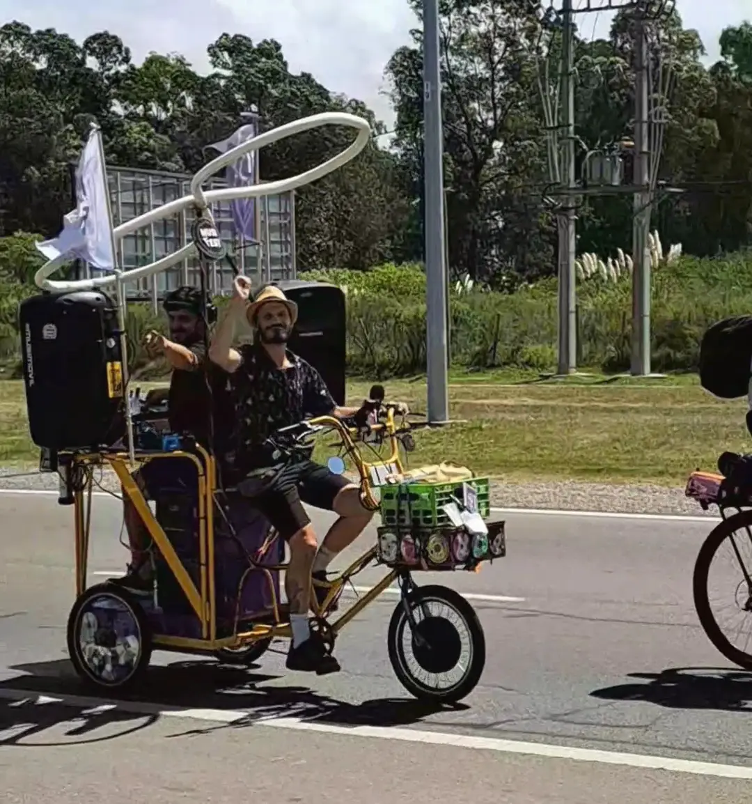 Pedro, in a straw hat and patterned shirt, pedals the tricycle along a wide avenue in daylight, a helmeted rider following close behind.