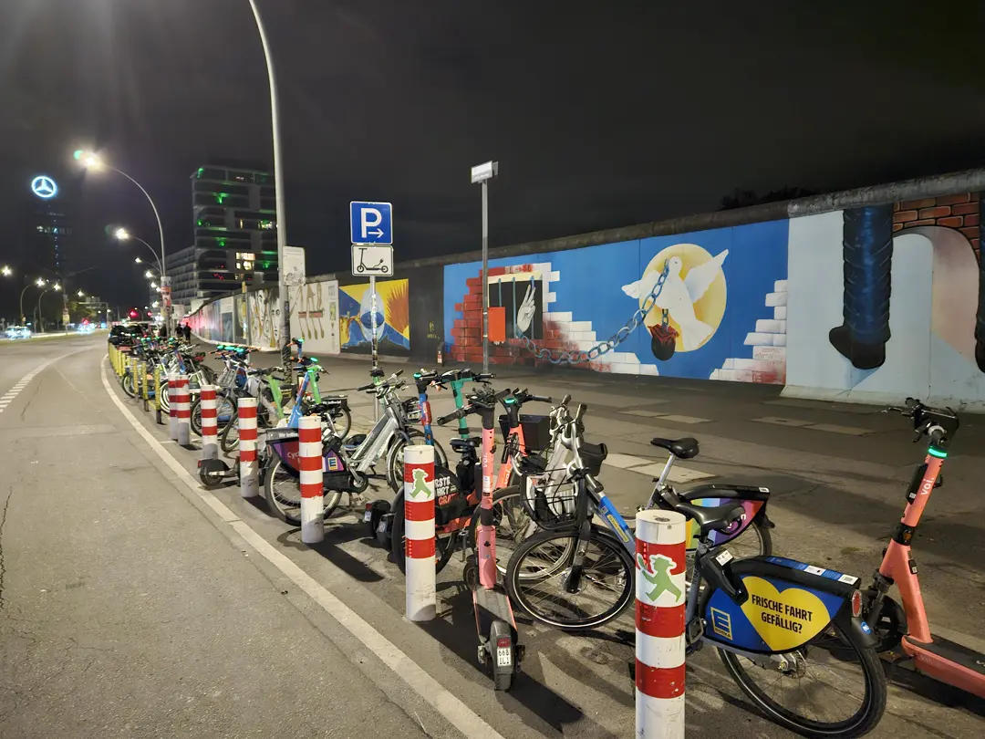 Bikes and scooters from several companies aligned in a marked bay beneath the East Side Gallery.
