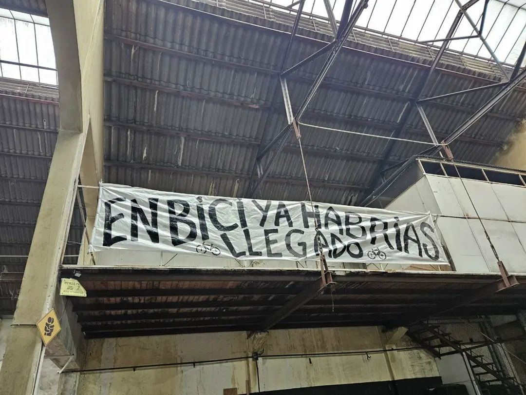 A hand-painted banner hanging from Rodala's mezzanine: «EN BICI YA HABRÍAS LLEGADO», under the warehouse's corrugated roof and steel trusses.