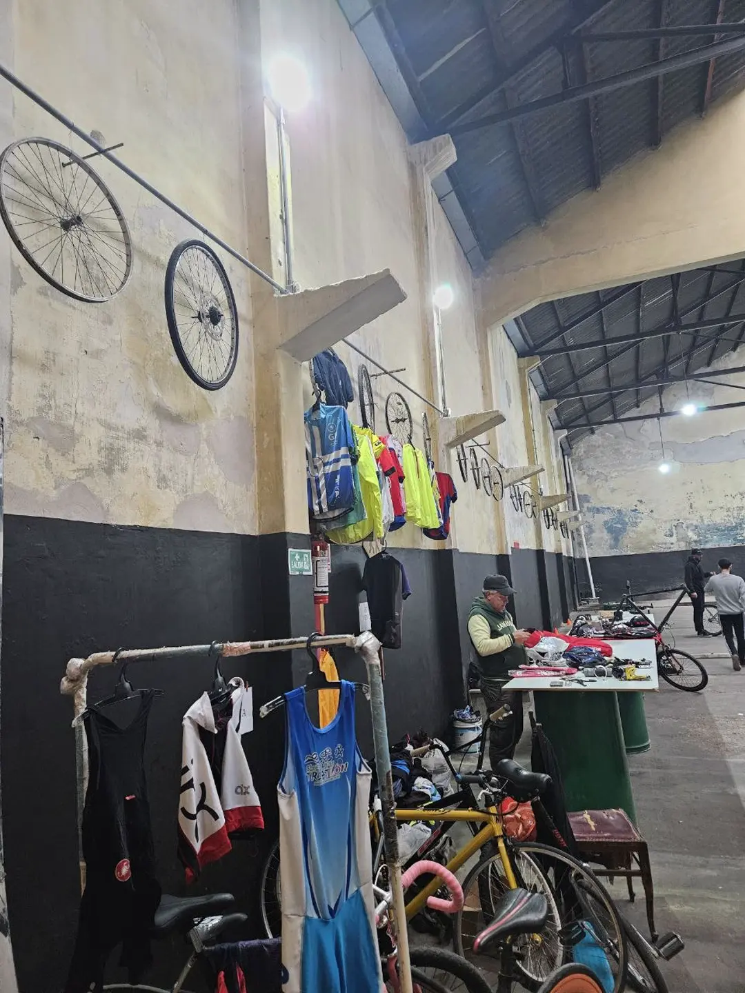 Stalls at Rodala's parts fair: bicycle wheels hung on the wall, second-hand jerseys on racks, and bikes leaned against a table of spare parts, under the warehouse's black-painted frieze.